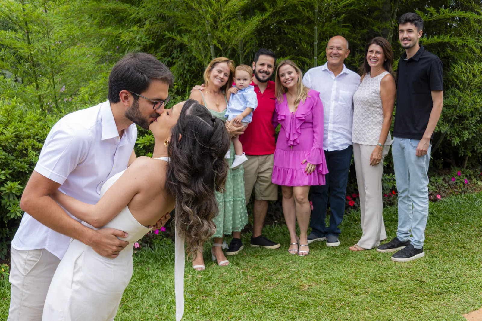 Amanda e Ricardo se beijando com familiares reunidos ao fundo no jardim